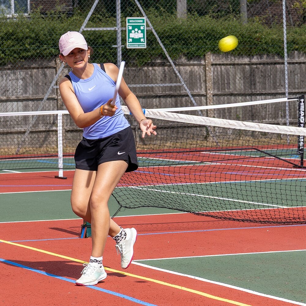 A student in a cap playing a shot at the net on an outdoor tennis court at Downe House
