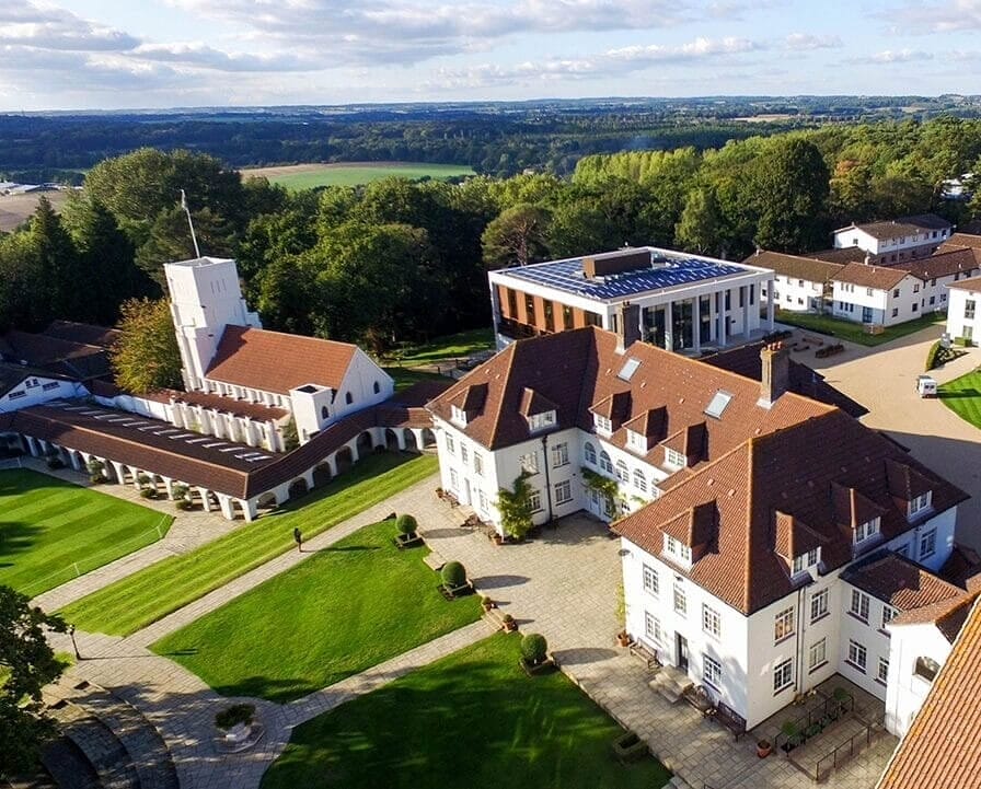 Aerial view of the Downe House campus: white buildings with red roofs around lawns, surrounded by woodland and open countryside