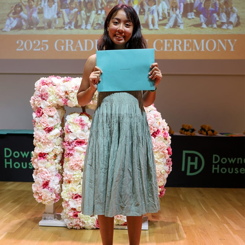 A summer school student holding her certificate on stage at the graduation ceremony, in front of a floral display with the school name