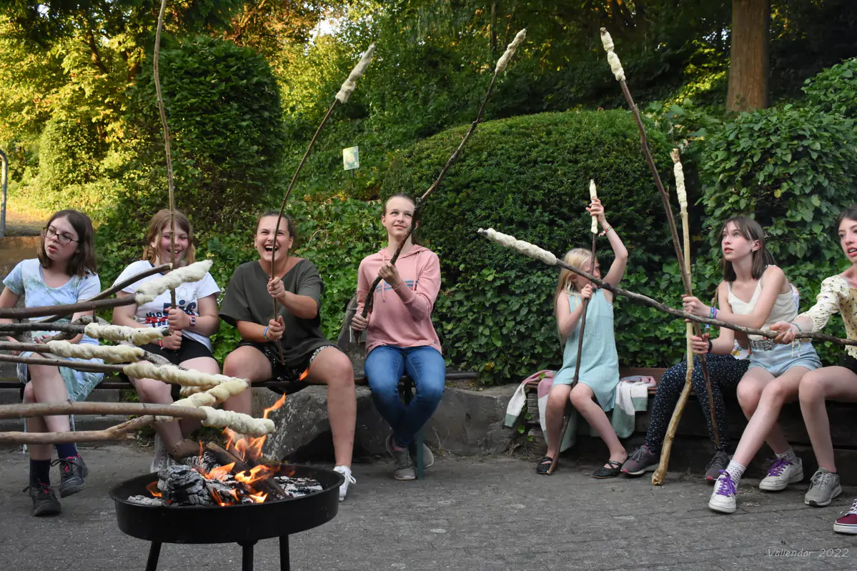 Children sitting around a fire bowl holding long sticks with bread dough, campfire evening at the Beginner Weeks camp in Vallendar