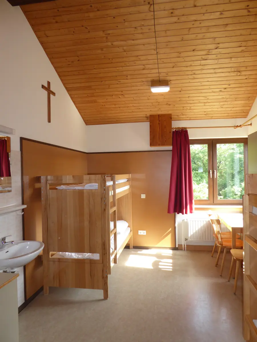 A bedroom with wooden bunk beds, a sloping pine ceiling, a washbasin and a window looking onto the grounds