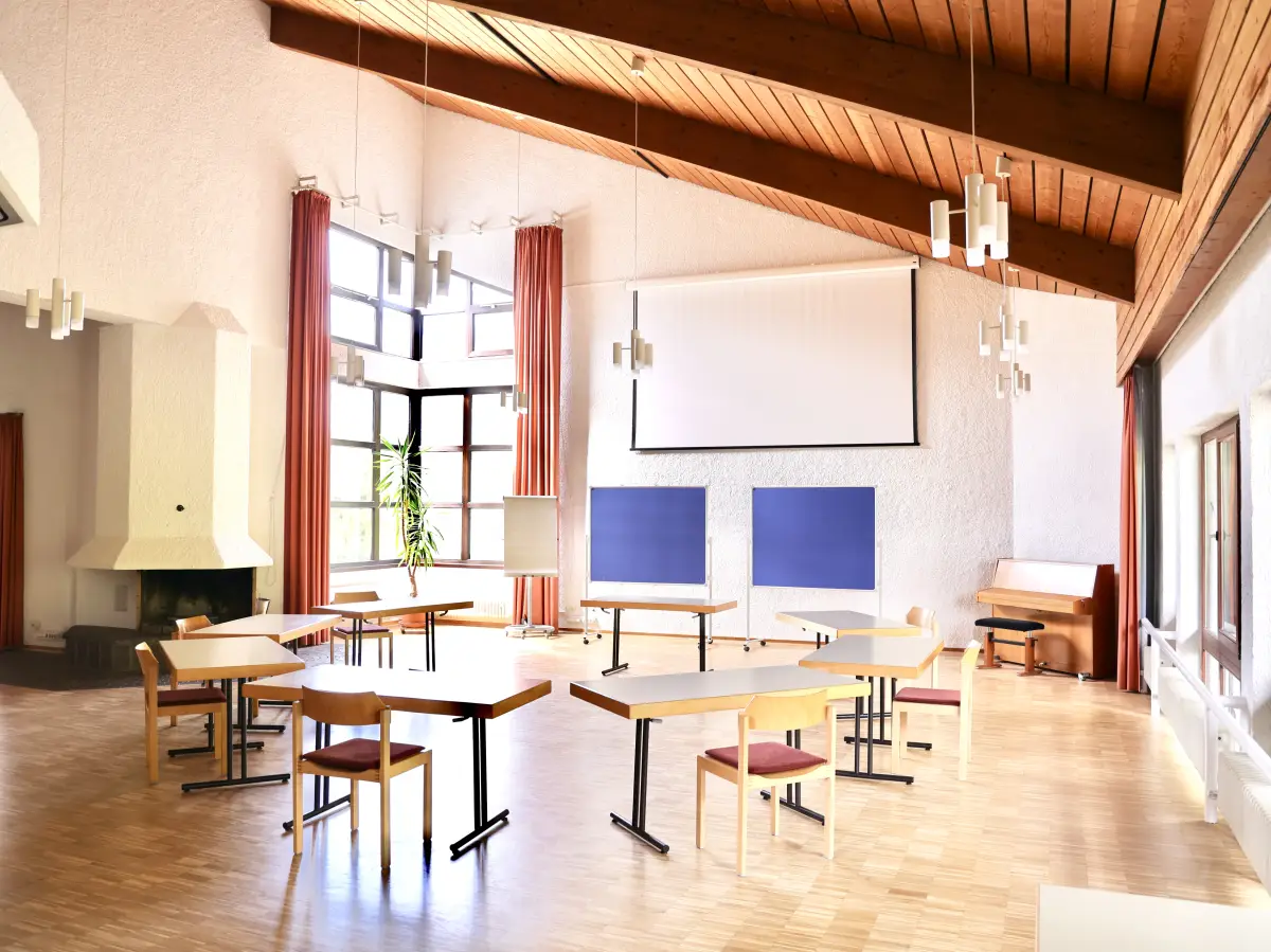 The large common room with a wooden ceiling, long tables and chairs, a projection screen, a piano and an open fireplace
