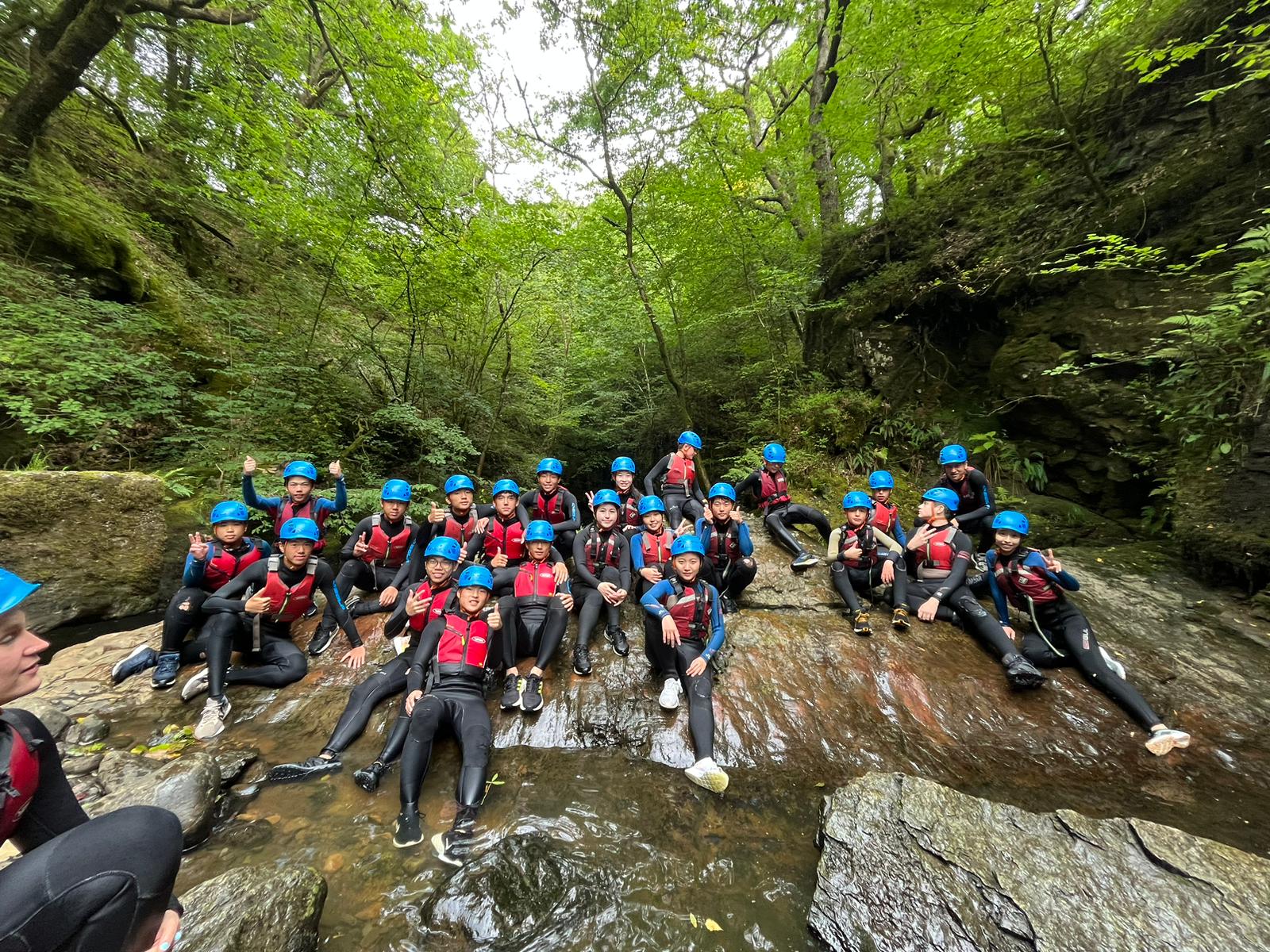 Large group of campers in wetsuits and helmets sitting on rocks in a wooded river gorge