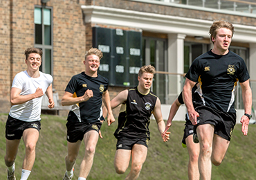 Four teenagers in black and white sports kit running together across a grass field beside a school building