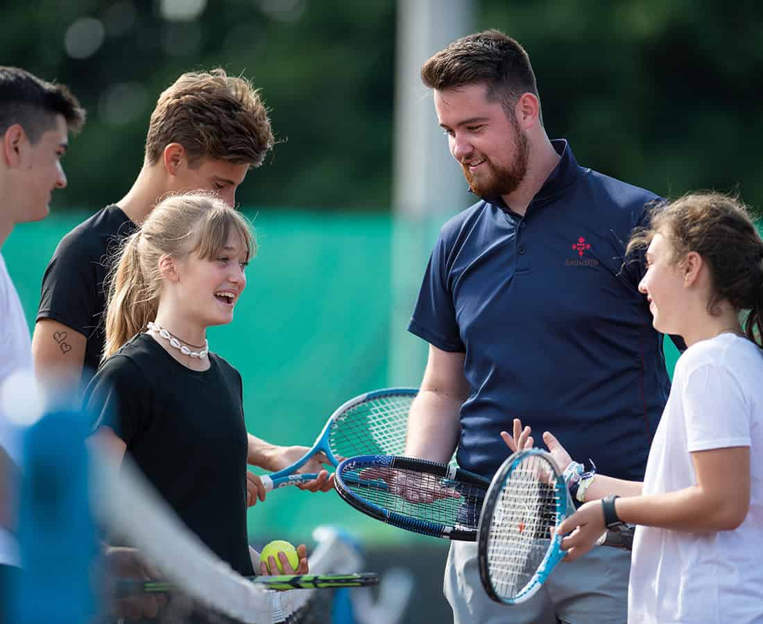A coach talks to students holding tennis rackets on an outdoor tennis court