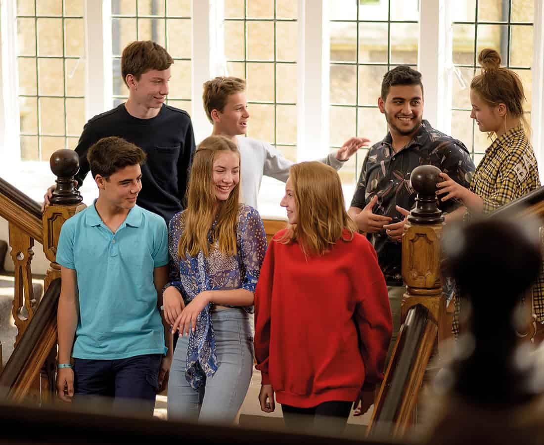 Students stand on a staircase inside a school building, talking and smiling together
