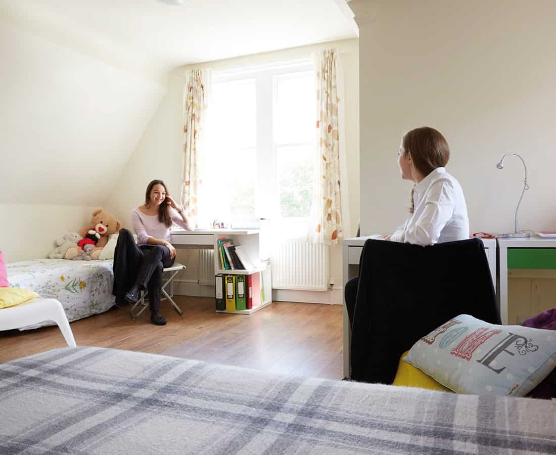 Two students sit and talk in a boarding bedroom with single beds and a window