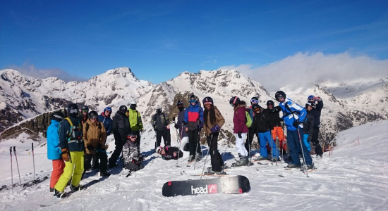 The whole group standing with skis and snowboards on a ridge with the snow-covered mountains behind