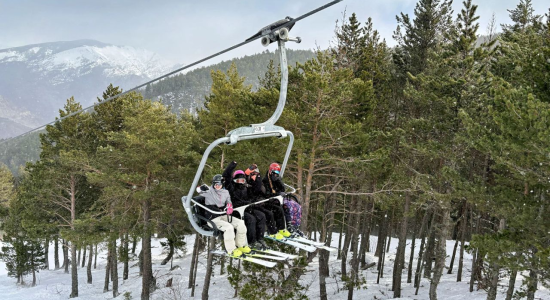 Four participants riding the chairlift above snow-covered pine trees