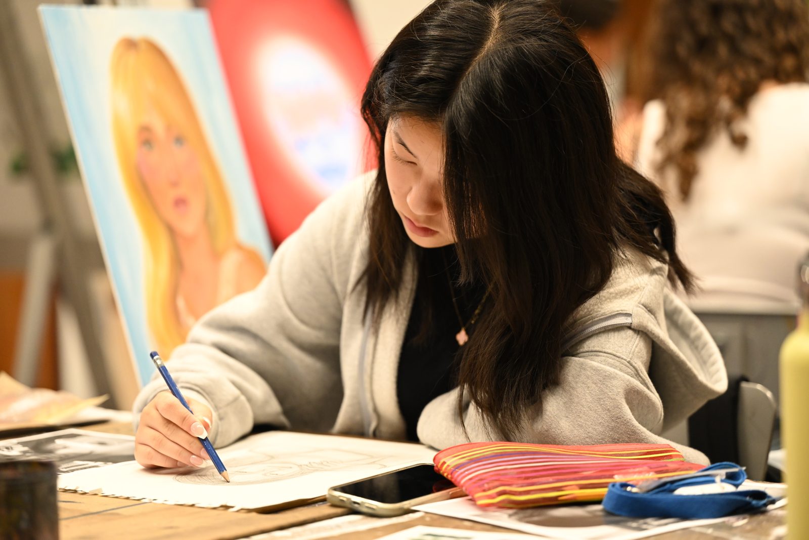 A student leans over their work with a pencil in hand at an easel in a bright studio, a painted portrait canvas standing beside them.