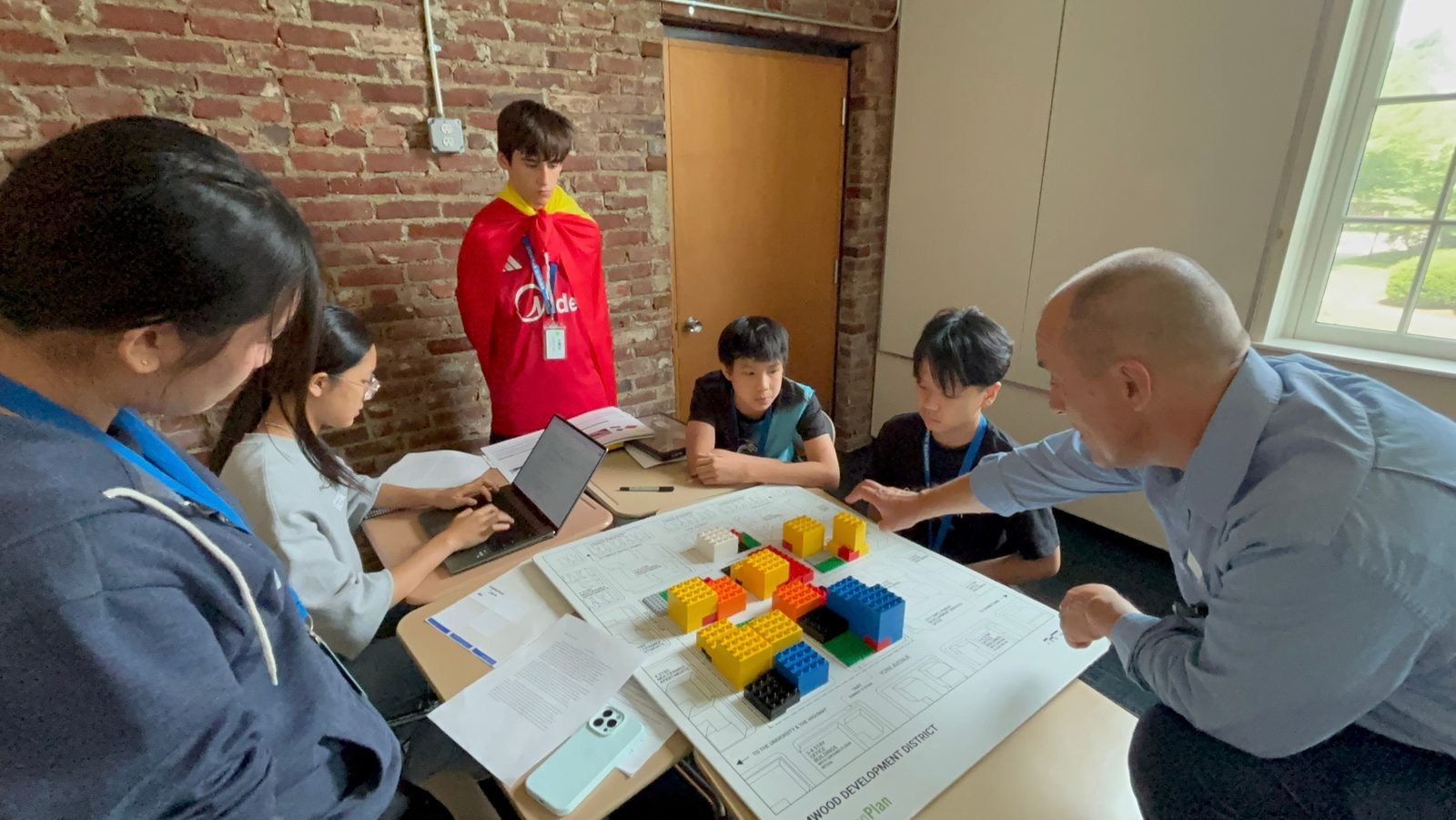 Students and a teacher gather round a table covered with small coloured blocks arranged into a model, in a classroom with an exposed brick wall and tall windows.