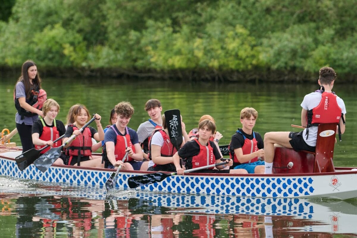 Pupils in red buoyancy aids paddling a dragon boat on the river with a member of staff steering