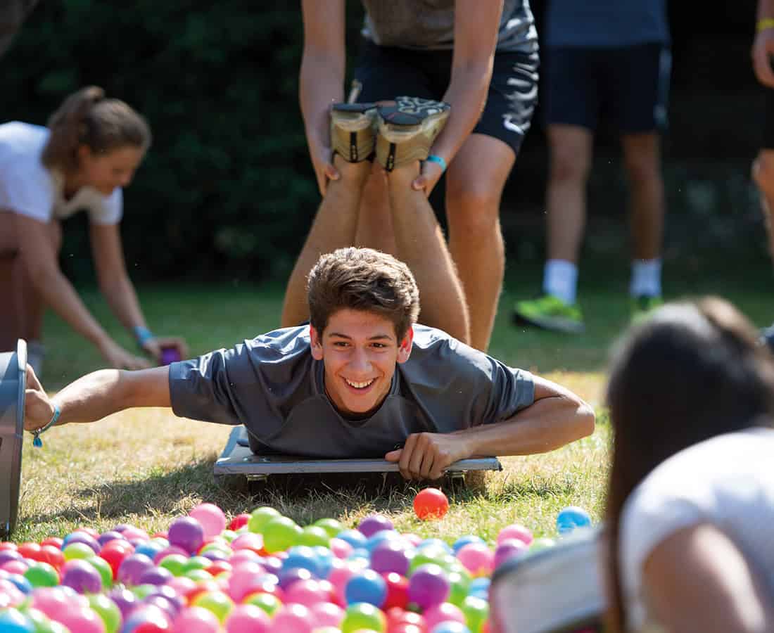 A student lying on a board in the campus garden during an outdoor game, classmates standing around him and coloured balls scattered on the grass