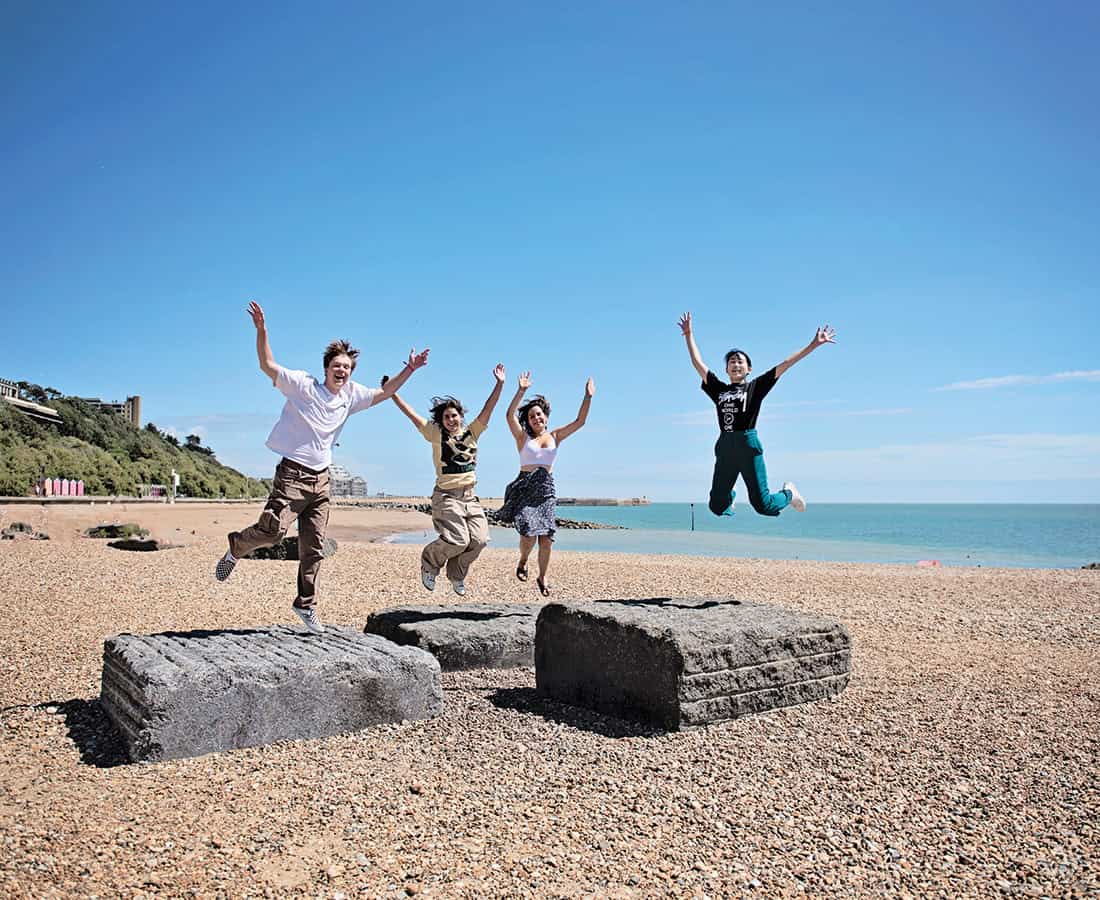 Four students jumping from low concrete blocks on the pebble beach at Folkestone, with the sea and the cliffs behind them