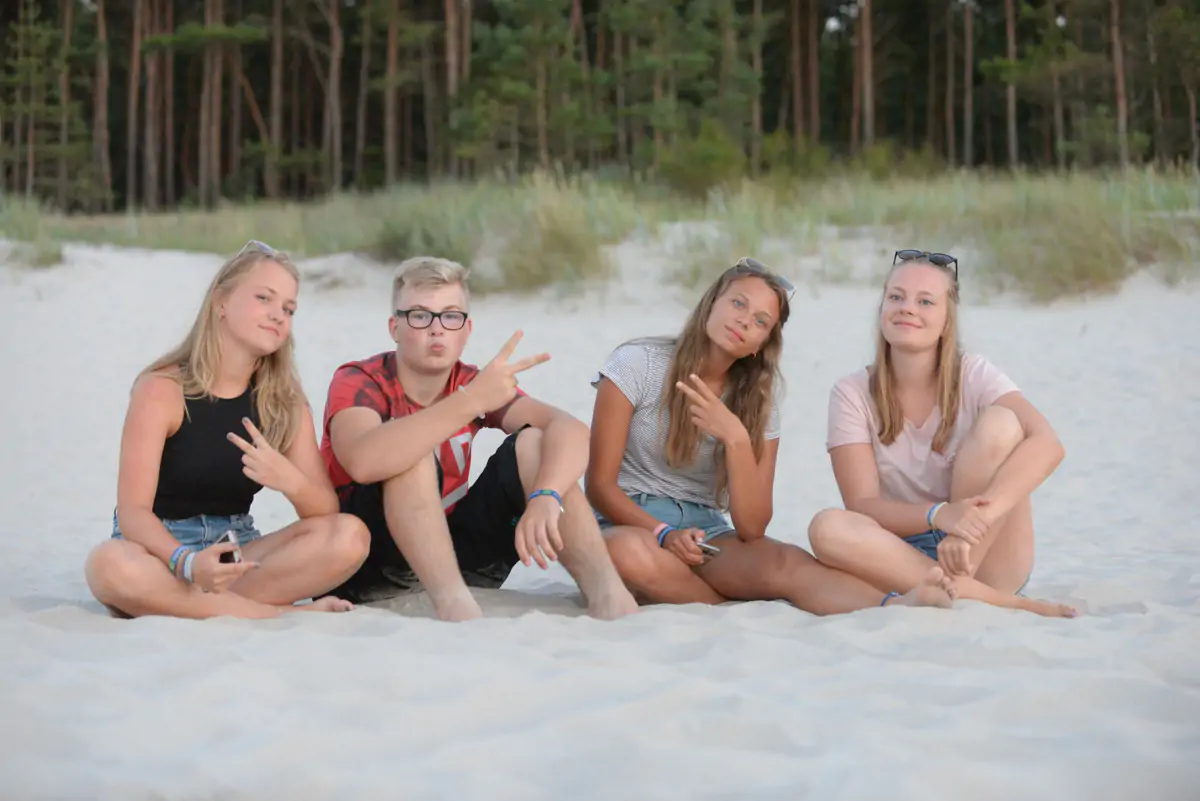 Four teenagers sitting on the white sand of the Baltic beach, with pine forest behind