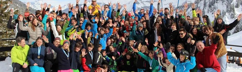Large group of people outdoors on snowy mountain, wearing winter clothes and raising hands, smiling for a group photo.