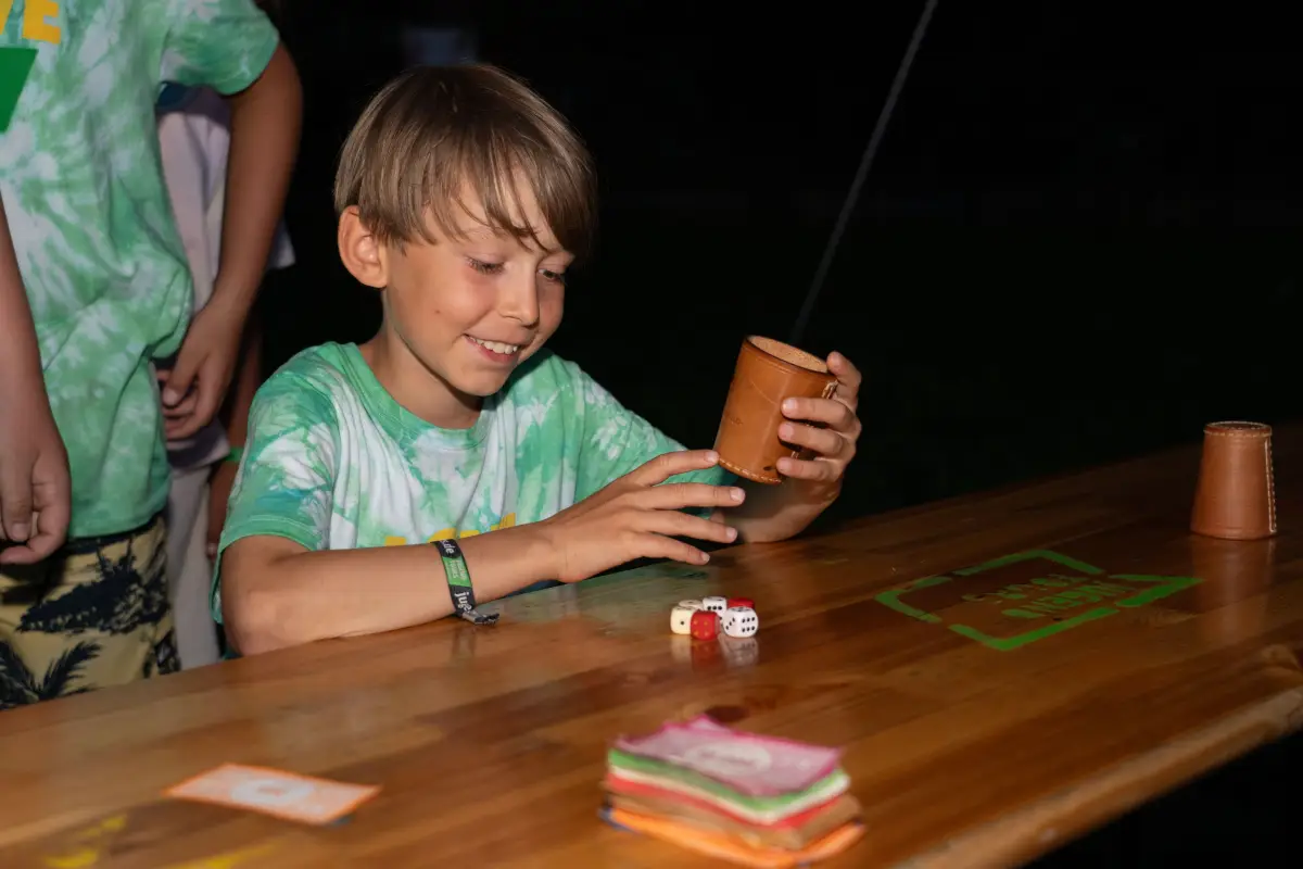 A boy playing a cup and dice game at a wooden table in the camp
