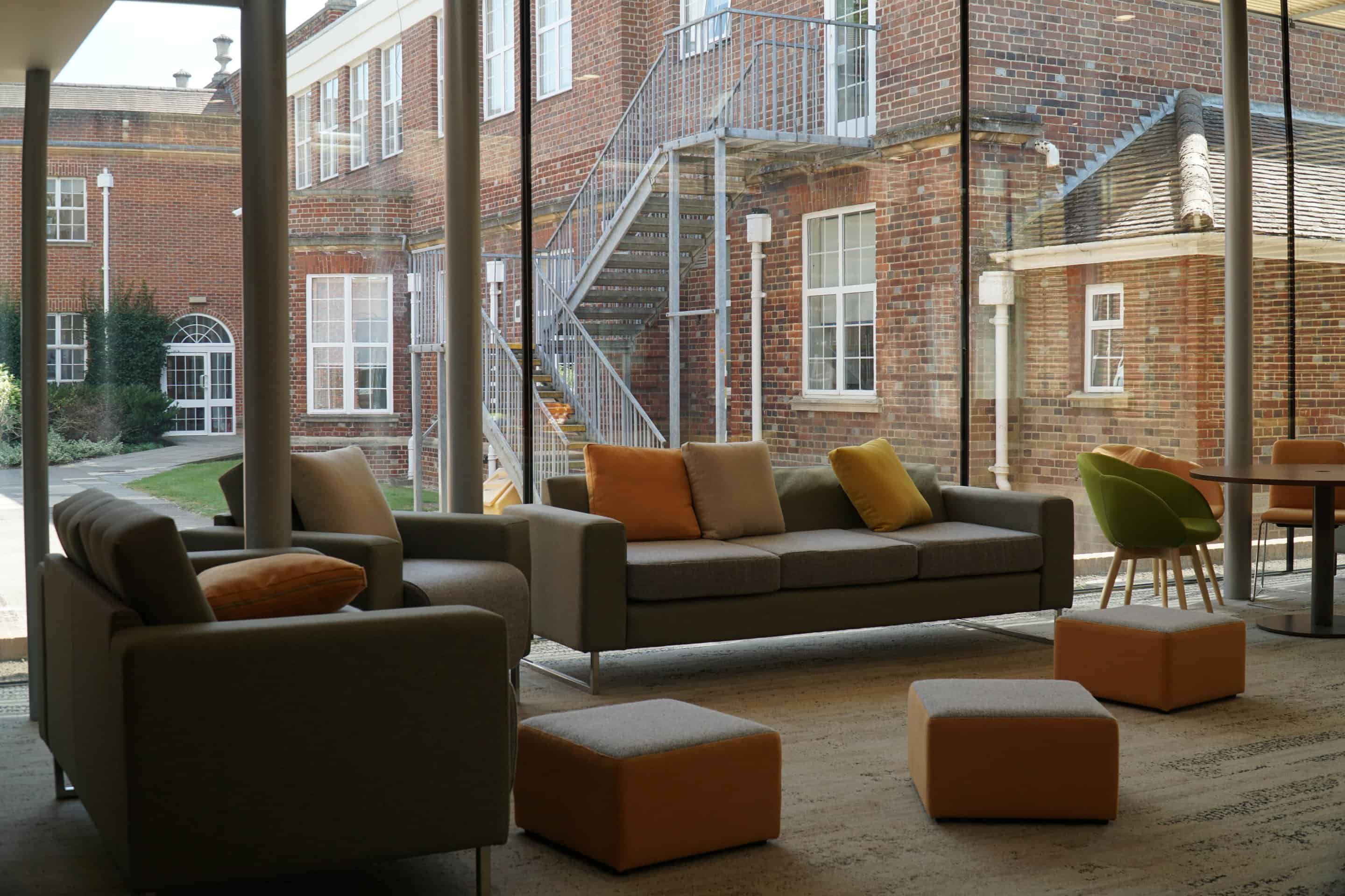 A common room with sofas and stools beside tall windows facing a brick courtyard