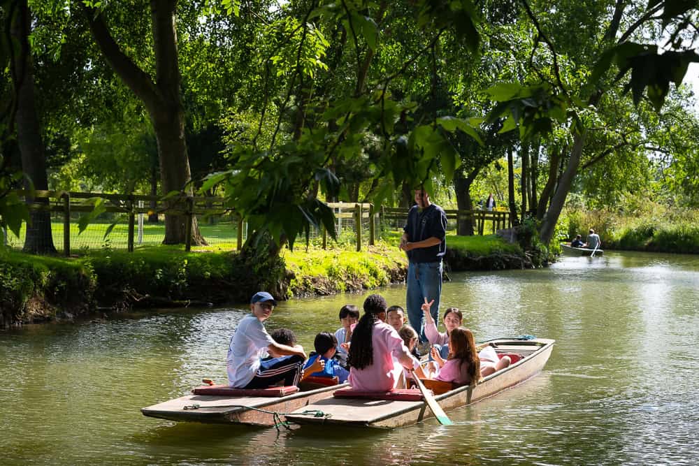 Students sit in punts on a tree lined river while an instructor uses the pole