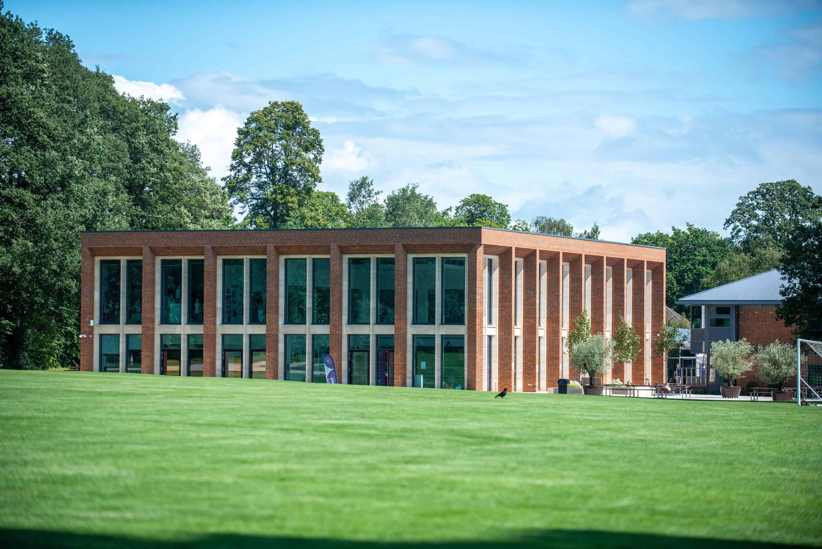 A modern hall with tall windows and columns stands beside a large green lawn