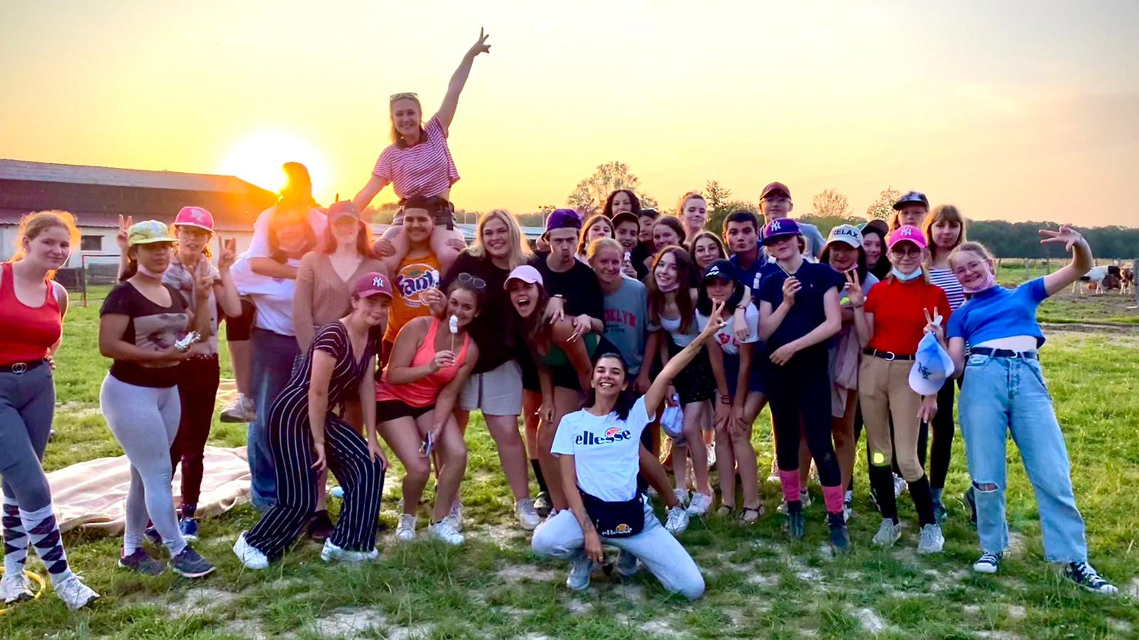 A large group of teenagers in riding clothes posing together in a field at sunset