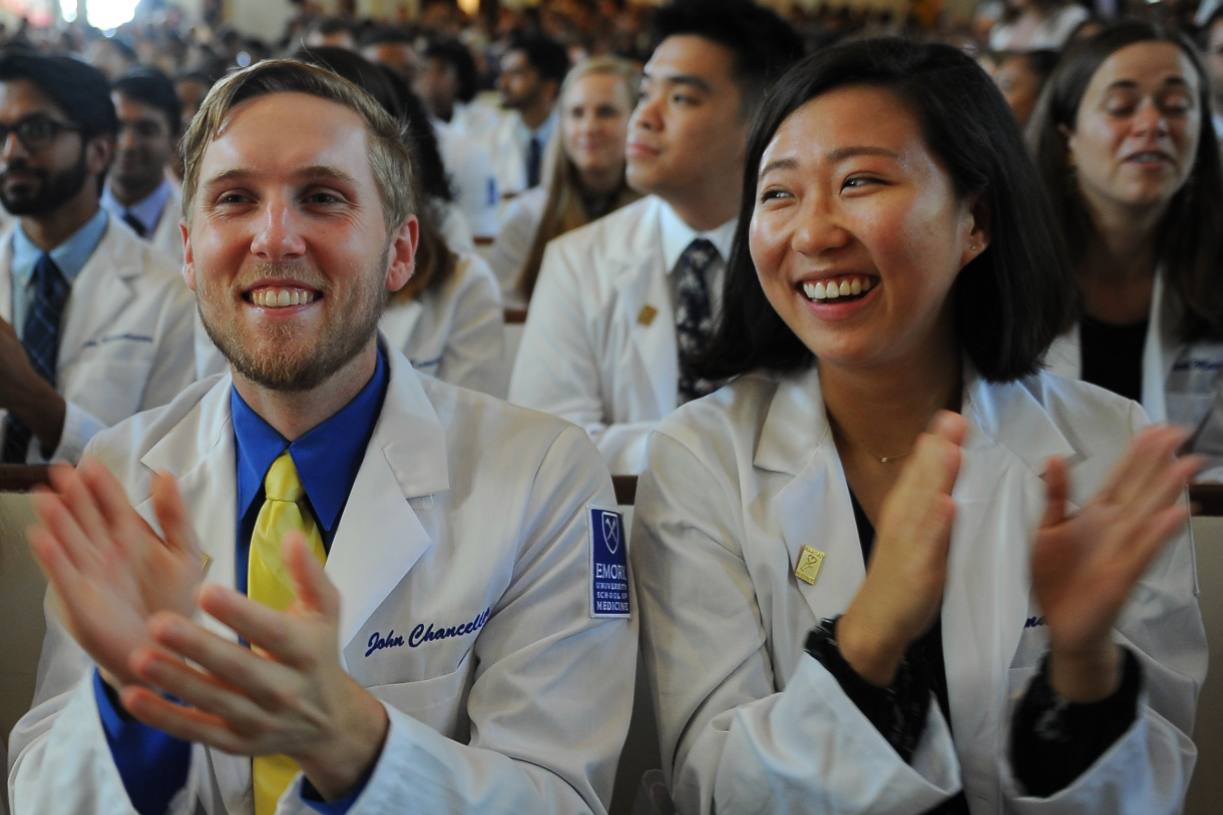 A packed auditorium of people in white coats applauding at an Emory white coat ceremony, the model for the ceremony that opens the Pre-Med Institute