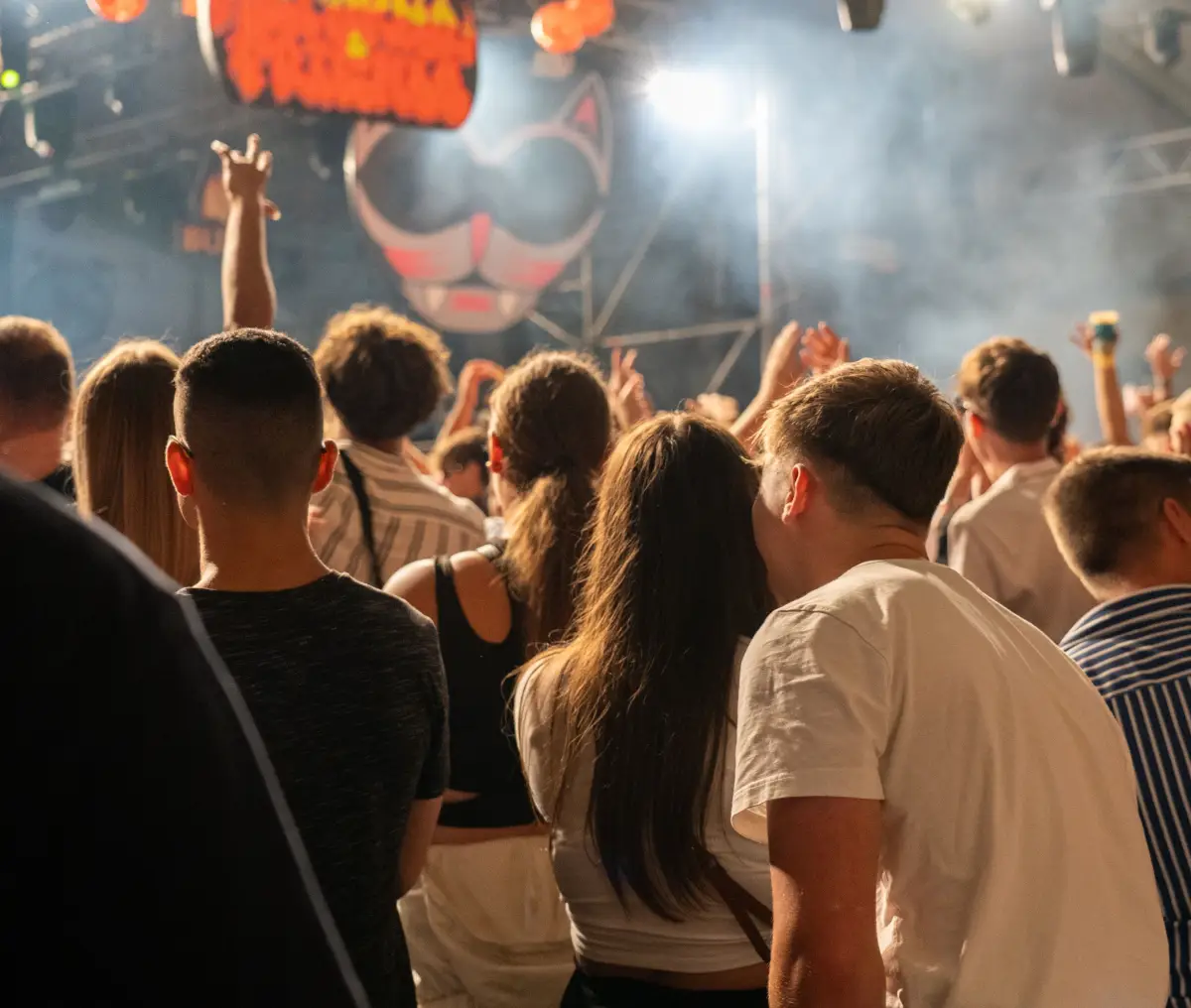 A crowd at an open-air party in front of a lit stage with smoke