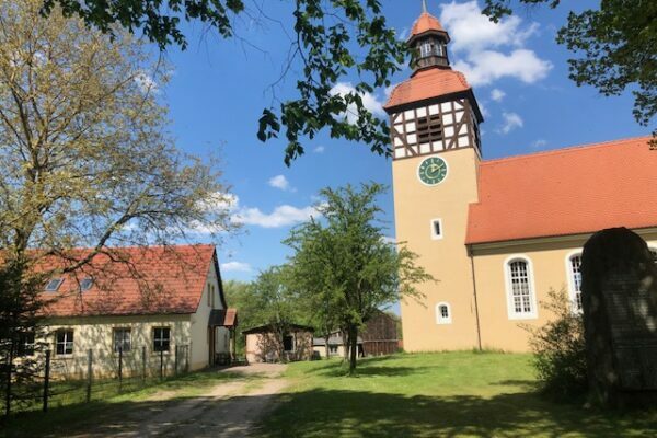 Village church with a half-timbered tower next to a red-roofed house in Vietmannsdorf, the setting of the Rüstzeitheim Schorfheide