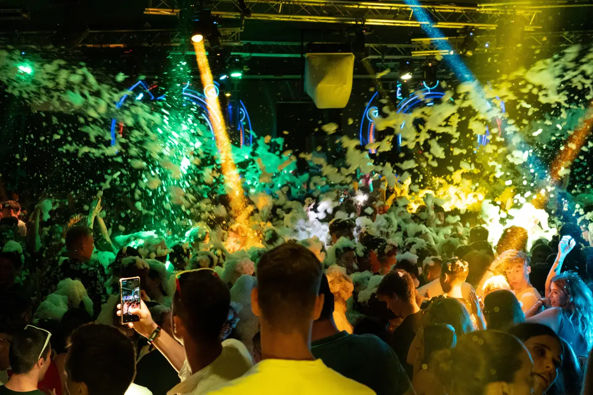 Crowded club night with green laser light and foam, participants filming on phones, during the Rimini evening programme