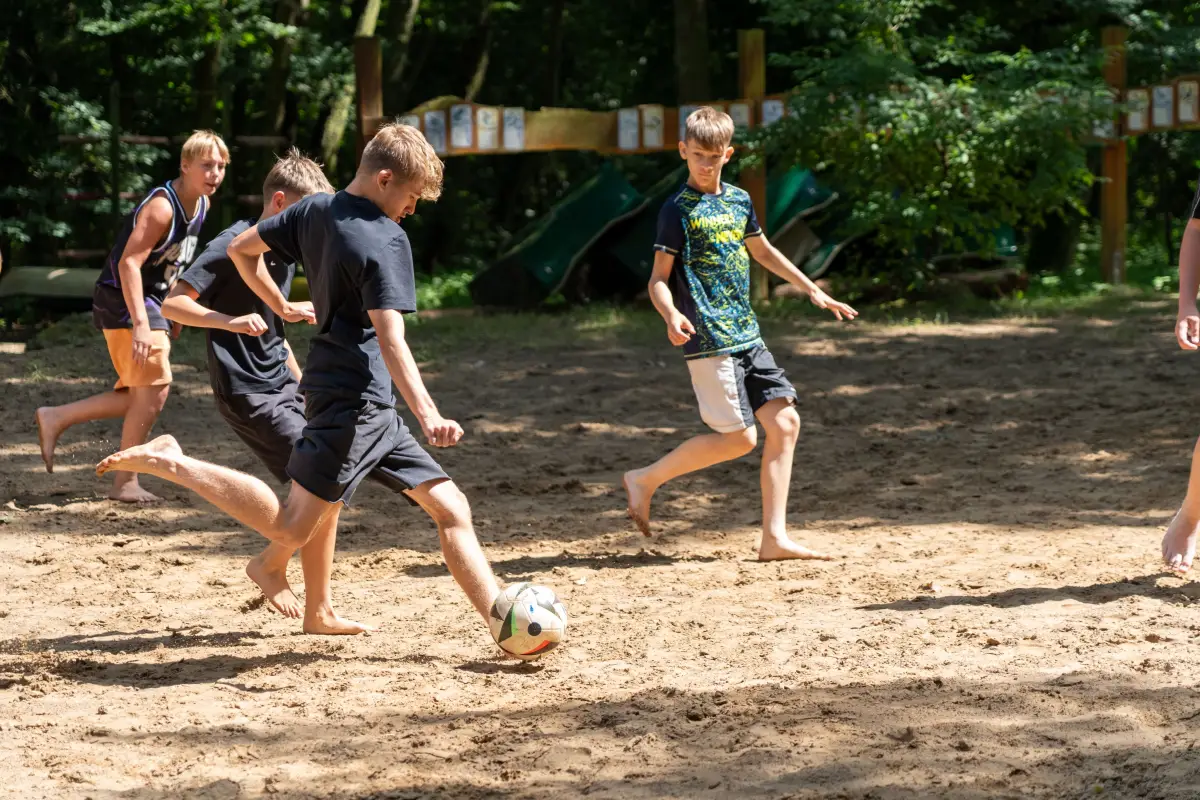 Campers playing football barefoot on the sandy ground beside the lake