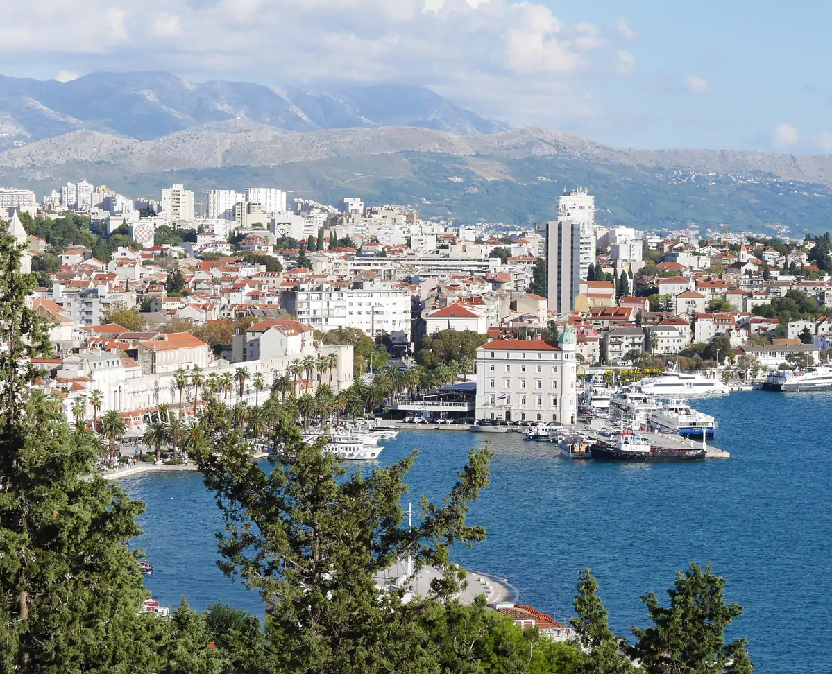 A harbour city of pale stone buildings along the water, with hills rising behind it