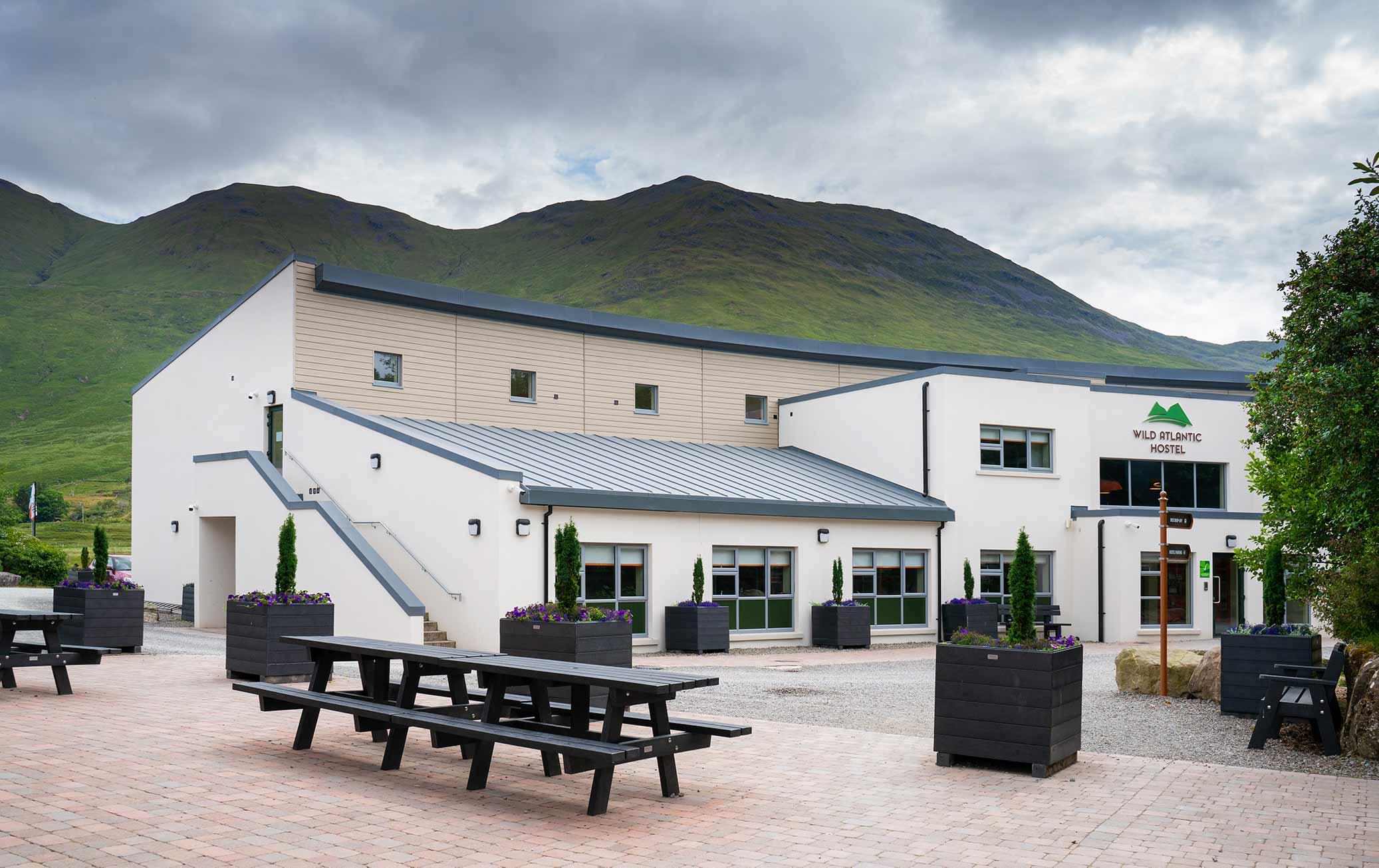 Exterior of the Wild Atlantic Hostel at Delphi Resort, with picnic benches outside and the Connemara mountains behind