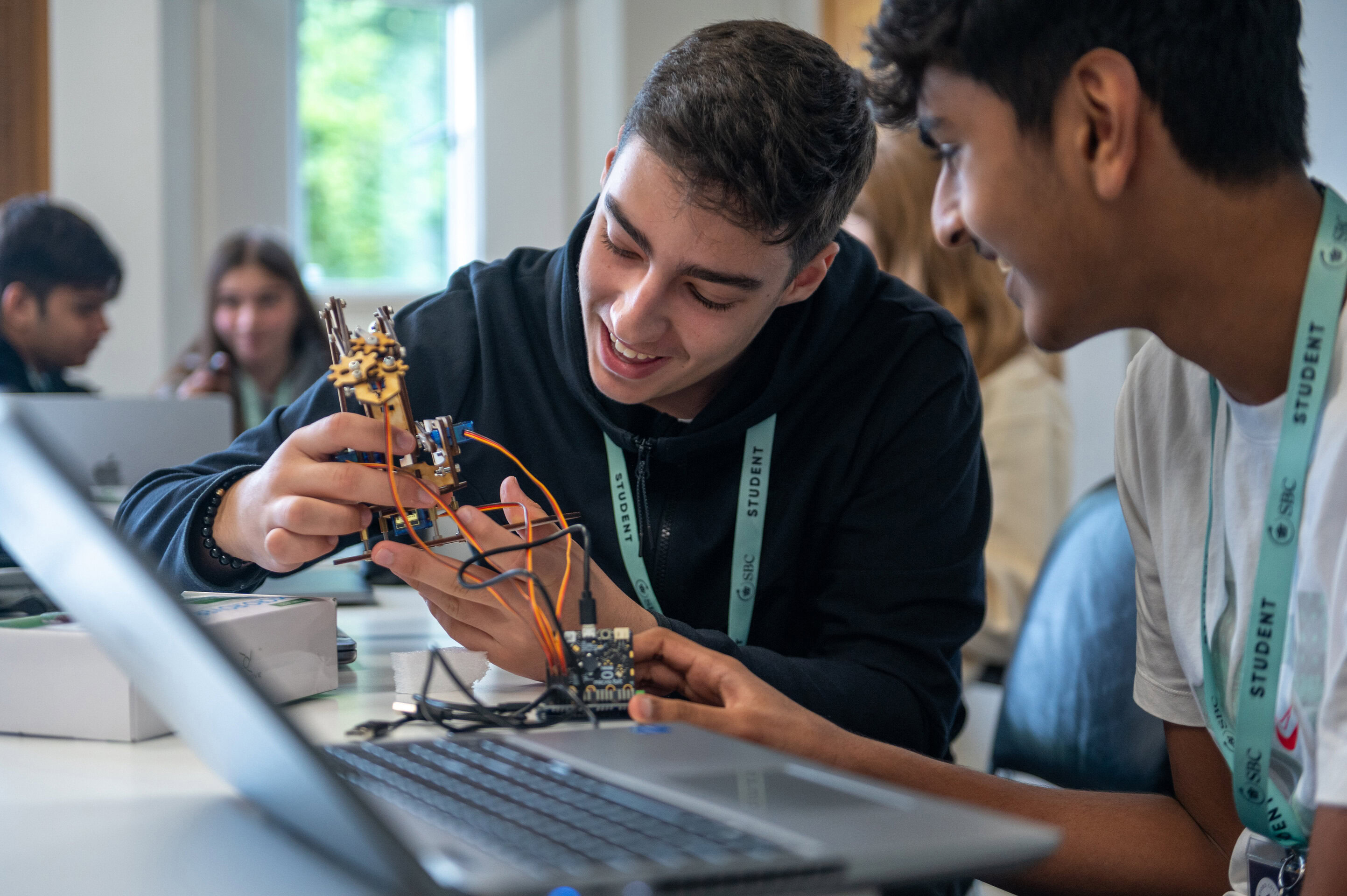 Two students look closely at a small robot beside a laptop in a classroom
