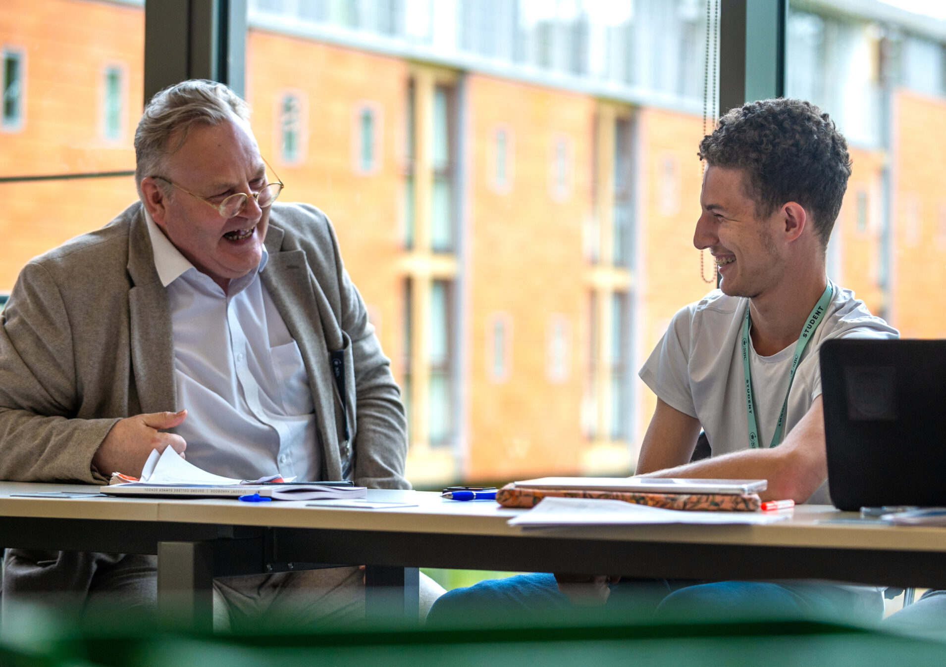 A tutor and a young man laugh together over papers at a table indoors