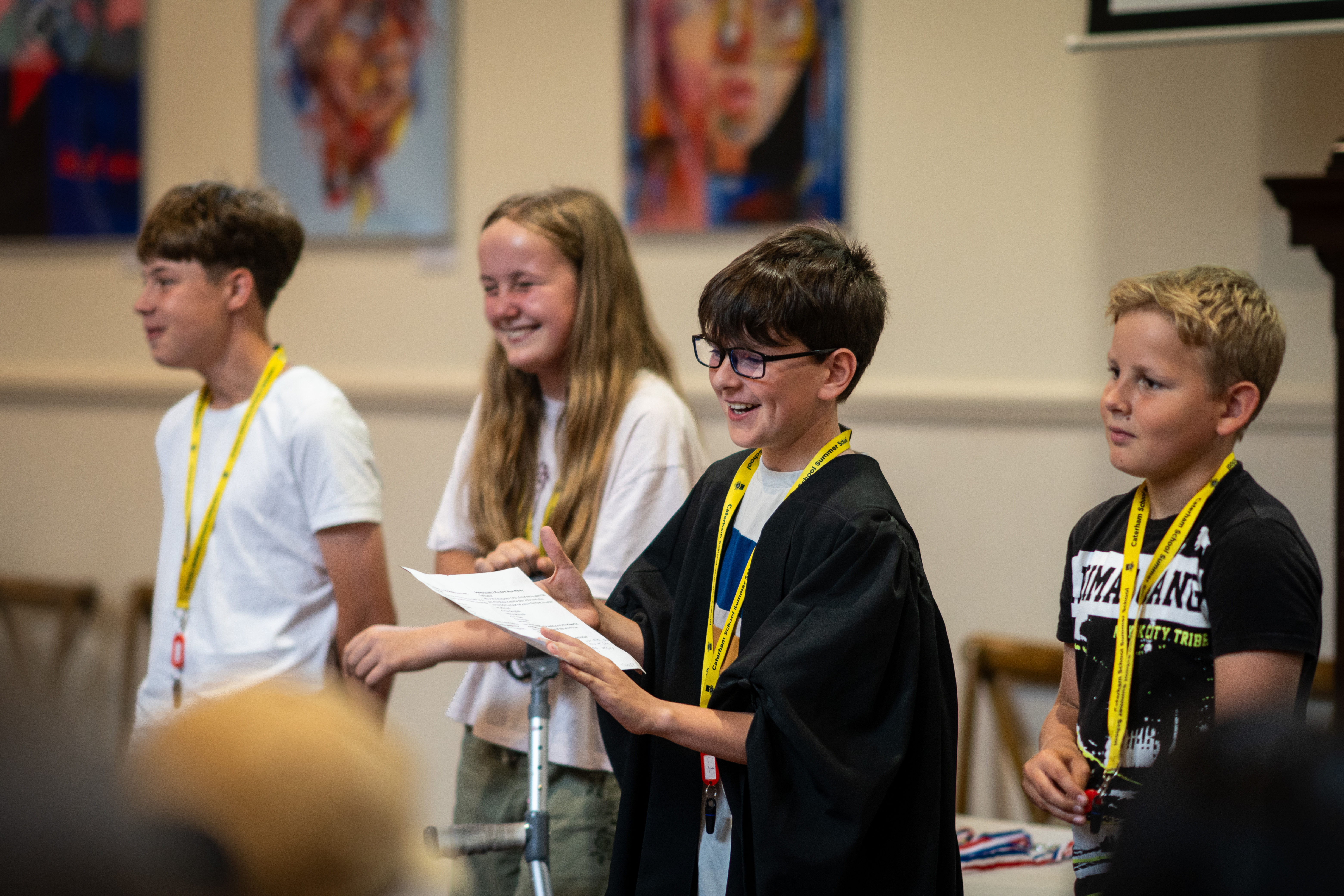Group of children in a classroom, one wearing a black academic gown, holding papers and speaking to the room