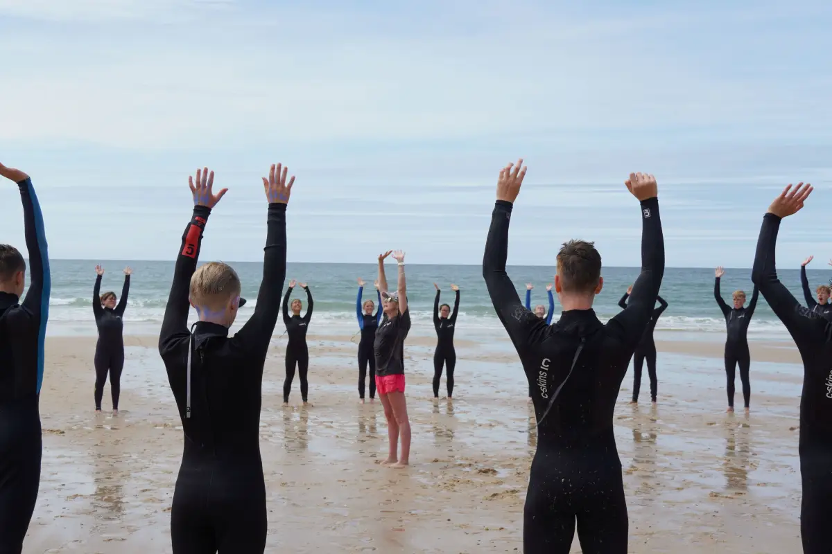 Participants in wetsuits warming up with arms raised on the beach before surfing