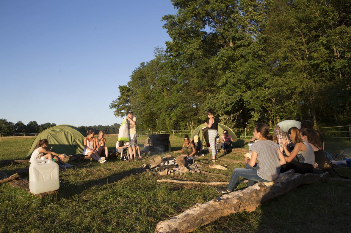 Campers sitting round a fire beside tents pitched in a meadow at dusk