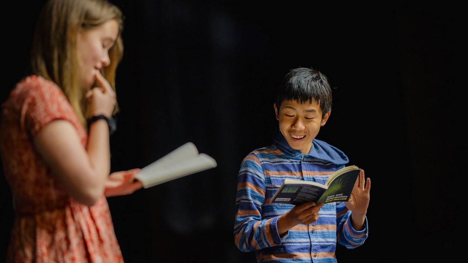 A student performing from a script under a spotlight on a darkened stage