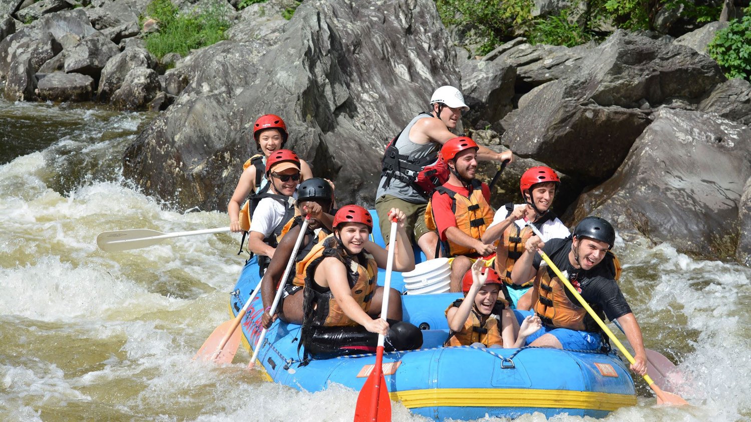 A raft of students in helmets paddling through whitewater on a river excursion