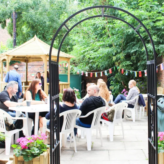 A photo of the garden and patio area to the rear of the school in ELC English language school in Chester. There is a metal archway you can walk through to get to a paved area with white patio seating and students are sitting chatting together. In the background is a wooden gazebo and lots of trees.