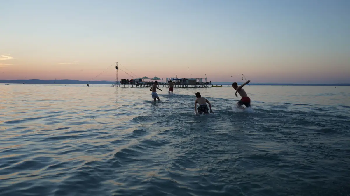 Swimmers standing and playing in the shallow water of Lake Balaton at sunset, with a wooden jetty on the horizon