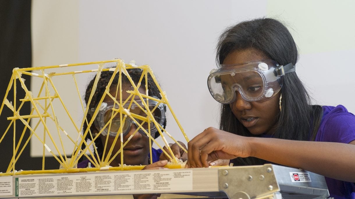 Two Engineering Innovation students in safety goggles setting up their spaghetti bridge for the competition