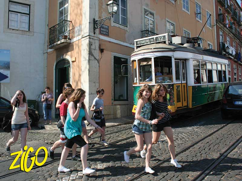 Yellow Lisbon tram coming up a narrow street past pastel buildings, young people on the pavement