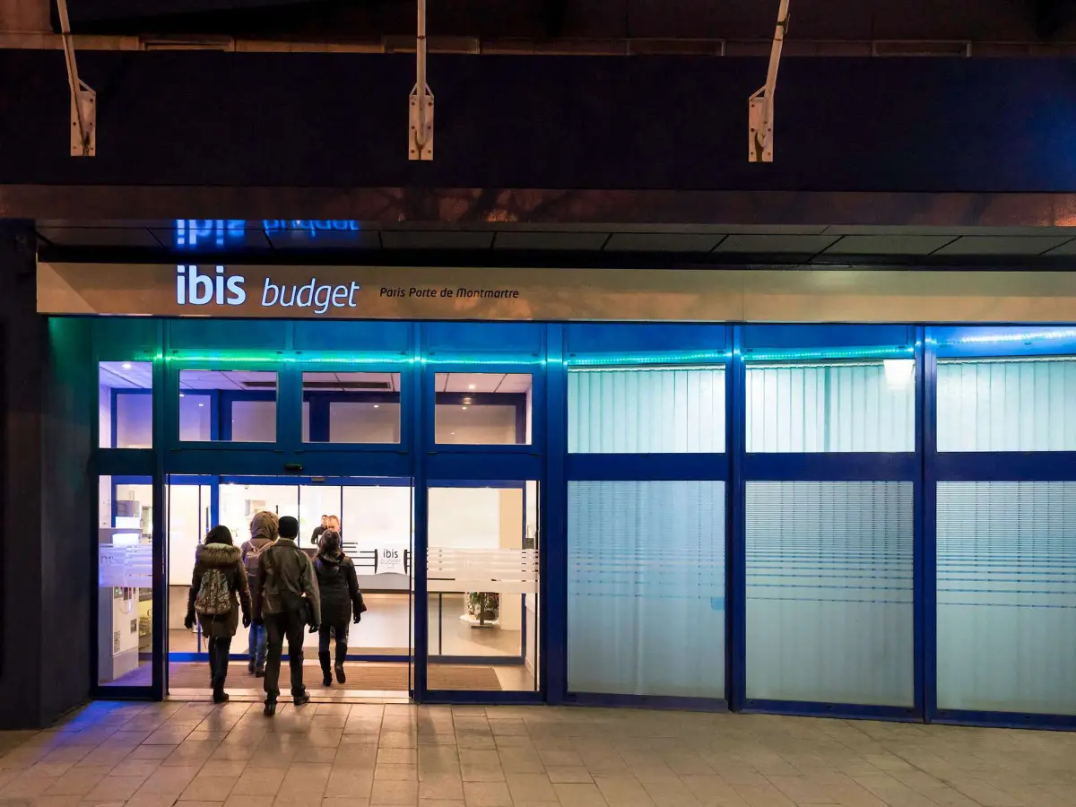 The lit glass frontage of an ibis budget hotel at night, with guests walking in
