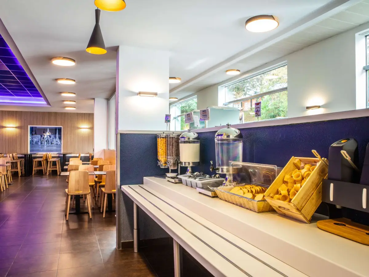 A hotel breakfast area with a buffet counter of cereal dispensers and fruit, and tables beyond