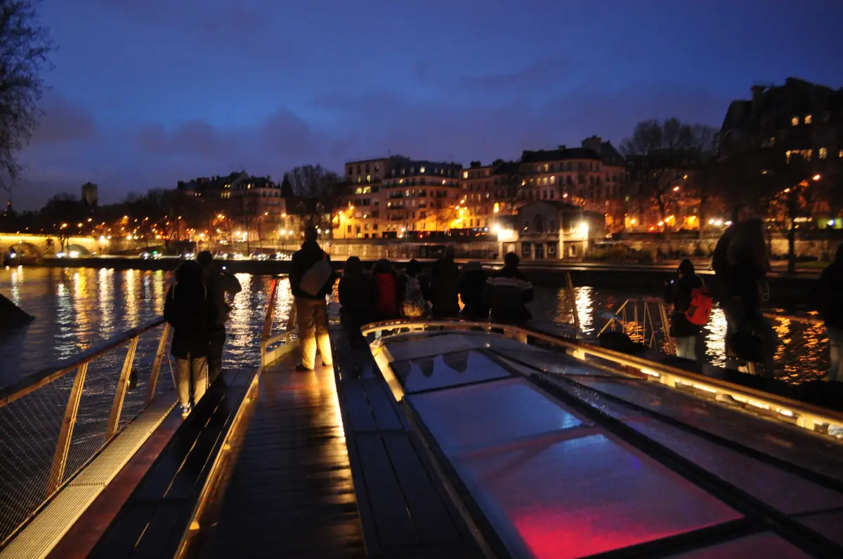 A glass-roofed sightseeing boat moored on the Seine at dusk, with lit buildings along the embankment
