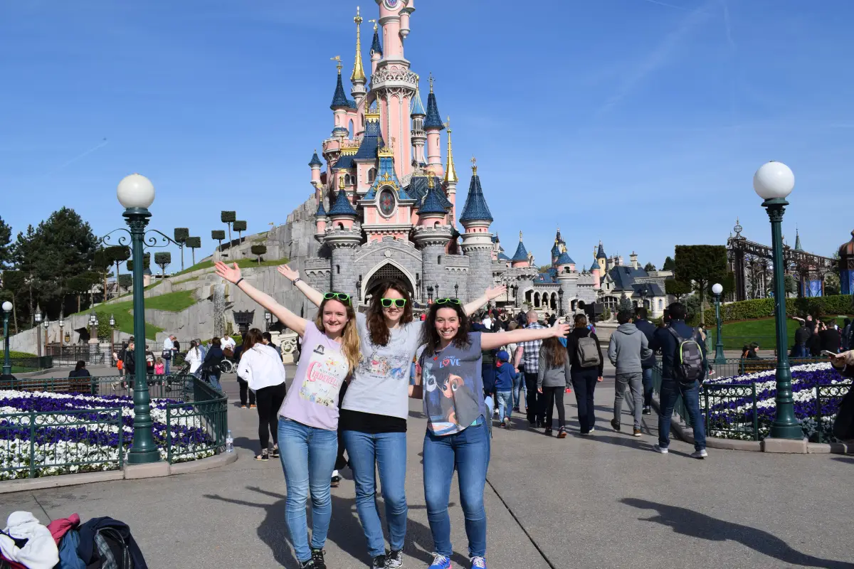 Three young travellers with arms raised in front of a fairy-tale castle, with visitors walking past