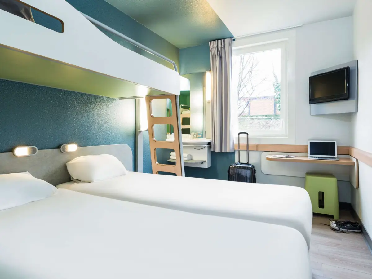 A compact hotel room with a double bed and a bunk above it, a wall-mounted television and a small desk