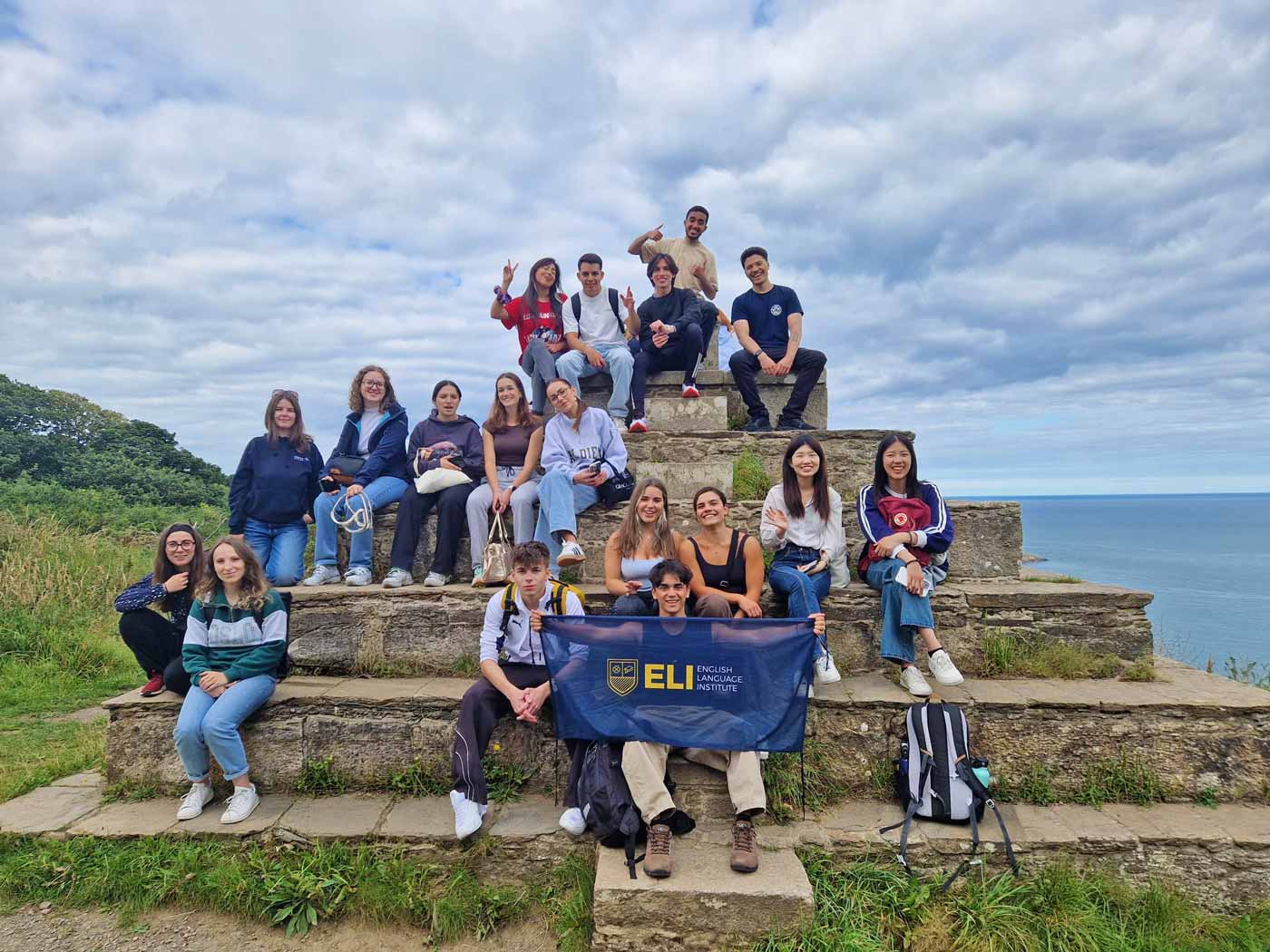 A large group of students sitting on stone steps on a clifftop above the sea with an ELI banner during an outdoor day