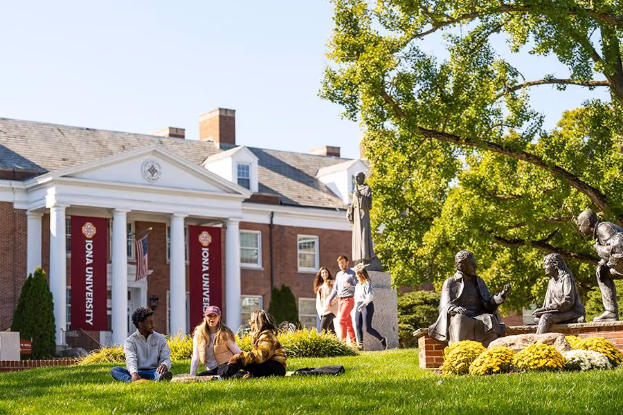 Students on the lawn in front of a colonnaded brick building with Iona University banners, New Rochelle campus